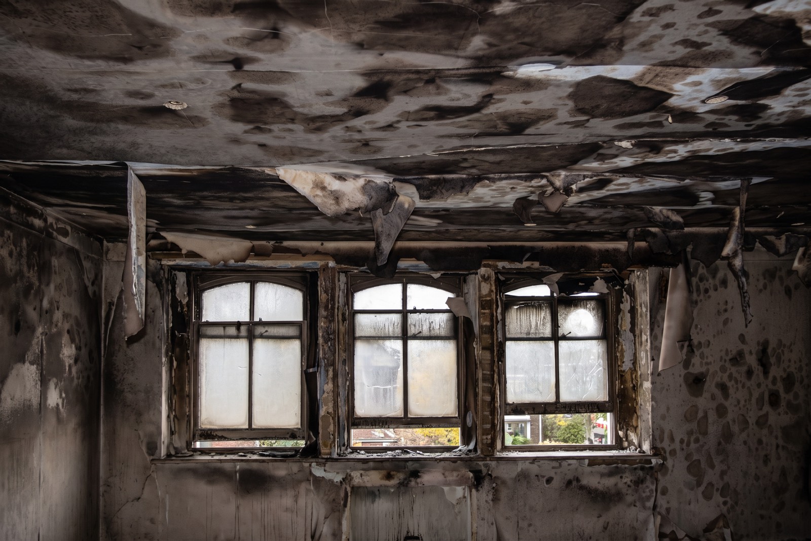 Fire-damaged room with a scorched ceiling, hanging ceiling lining and smoke-blackened walls around three windows